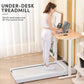 White under-desk treadmill with woman in white athletic wear standing on it while working at wooden desk with laptop