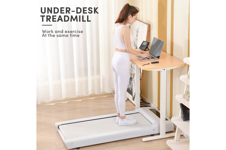 White under-desk treadmill with woman in white athletic wear standing on it while working at wooden desk with laptop
