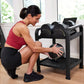 Woman in red tank top and black leggings selecting adjustable dumbbell from black Matrix dumbbell rack in home gym setting