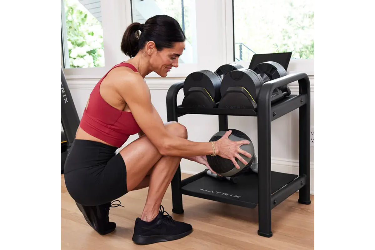 Woman in red tank top and black leggings selecting adjustable dumbbell from black Matrix dumbbell rack in home gym setting