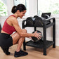 Woman in burgundy sports bra and black leggings selecting dumbbell from black metal storage rack in home gym