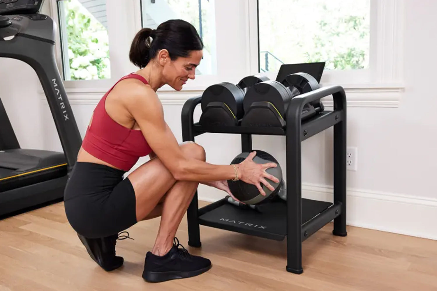 Woman in burgundy sports bra and black leggings selecting dumbbell from black metal storage rack in home gym