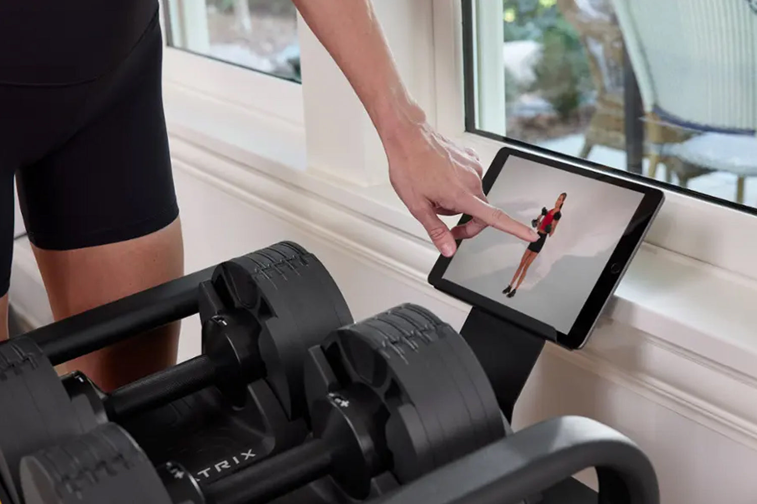 Person viewing fitness workout on tablet next to black adjustable dumbbells on windowsill