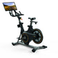Black stationary exercise bike with orange accents, tablet console mounted on handlebar displaying road scenery, on white bac