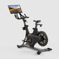 Black and gold stationary exercise bike with interactive monitor displaying desert road scene, on white background