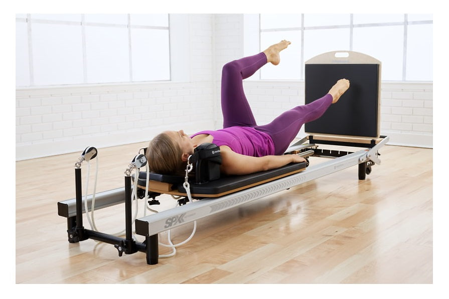 Woman in purple leggings performing leg exercise on white and black Pilates reformer with wooden footbar, indoors