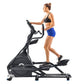 Woman using black Nautilus E618 elliptical machine, wearing black sports bra and blue shorts on white background