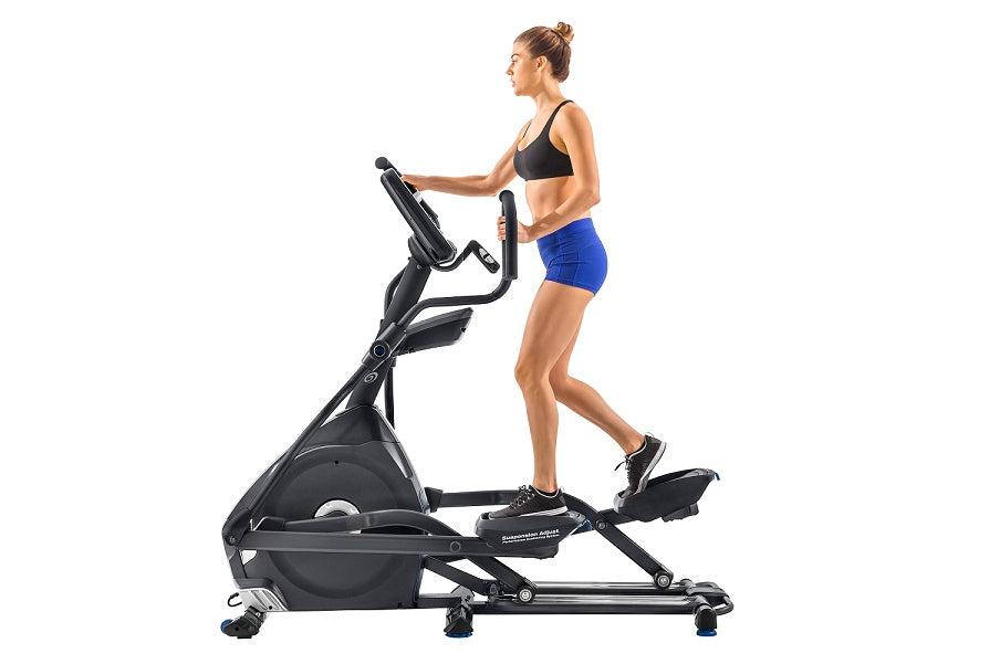 Woman using black Nautilus E618 elliptical machine, wearing black sports bra and blue shorts on white background