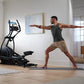 Man in gray tank top and tan shorts performing yoga pose next to black NordicTrack AirGlide 16 elliptical in modern home gym 