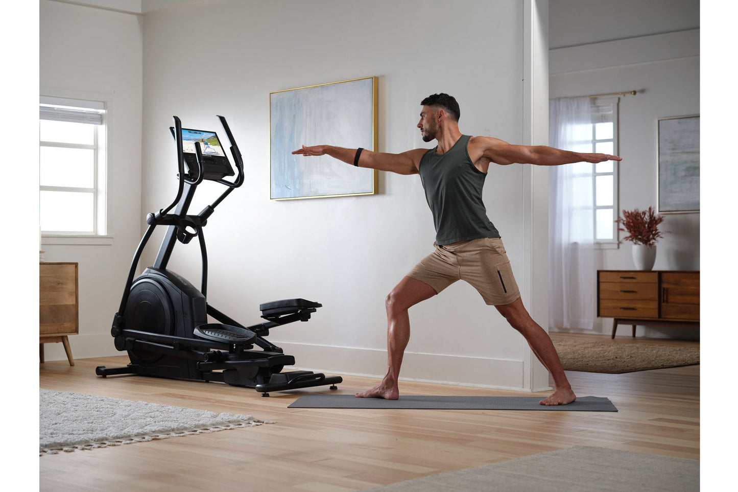 Man in gray tank top and tan shorts performing yoga pose next to black NordicTrack AirGlide 16 elliptical in modern home gym 
