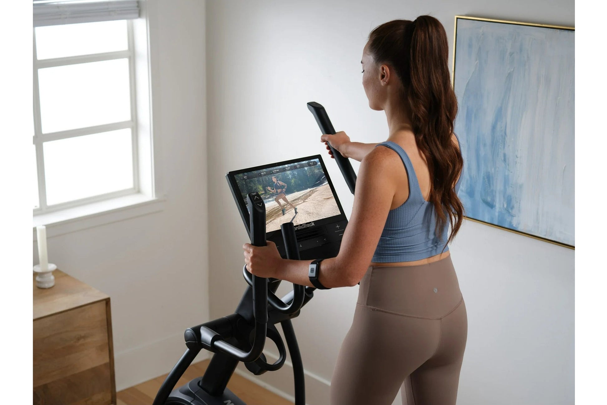 Woman wearing blue sports bra and tan leggings using black NordicTrack AirGlide 16 elliptical with digital display in home gy