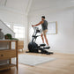 Man wearing charcoal tank top and tan shorts using black elliptical machine in bright white room with wooden furniture