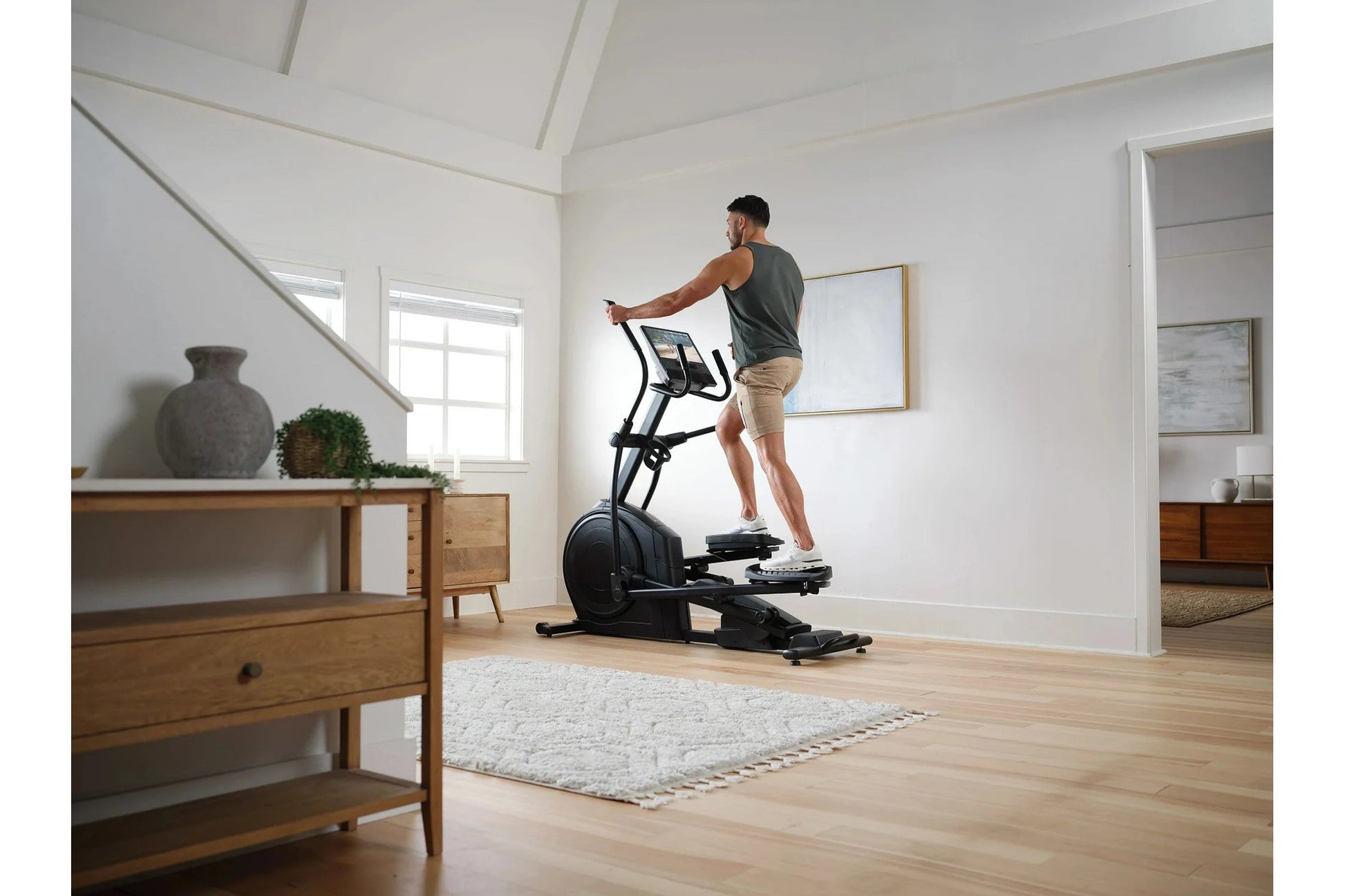 Man wearing charcoal tank top and tan shorts using black elliptical machine in bright white room with wooden furniture