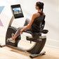 Woman seated on black and gray recumbent exercise bike with digital screen display, worn in home gym setting
