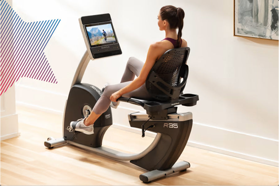 Woman seated on black and gray recumbent exercise bike with digital screen display, worn in home gym setting