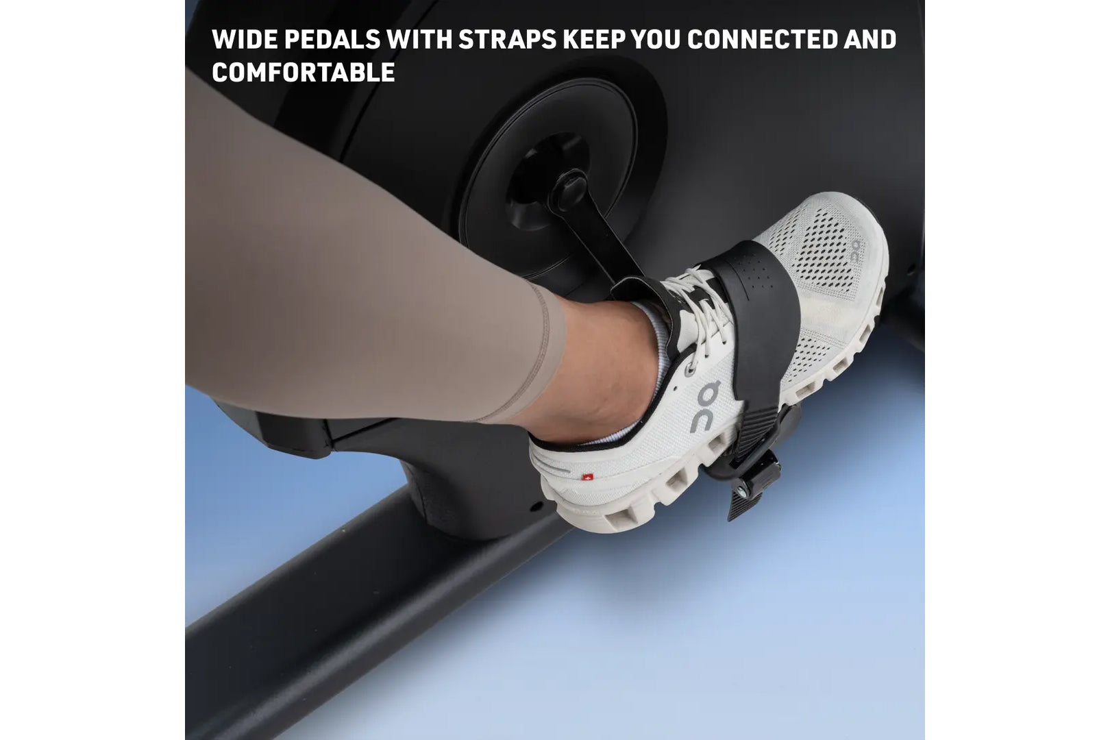 Foot on white and black exercise bike pedal with black strap, worn by model in beige leggings