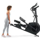 Woman in light gray leggings and white sneakers standing beside black NordicTrack AirGlide 14i elliptical machine on white ba