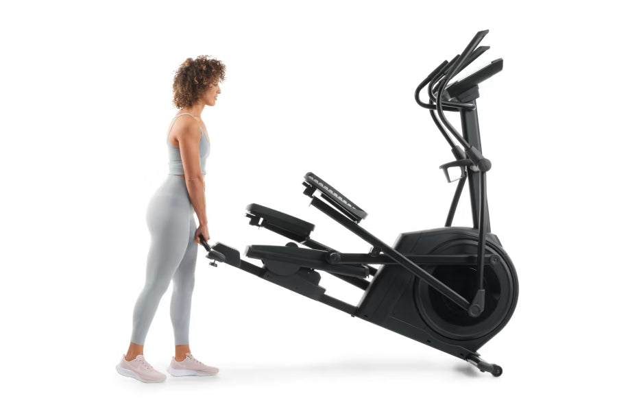 Woman in light gray leggings and white sneakers standing beside black NordicTrack AirGlide 14i elliptical machine on white ba