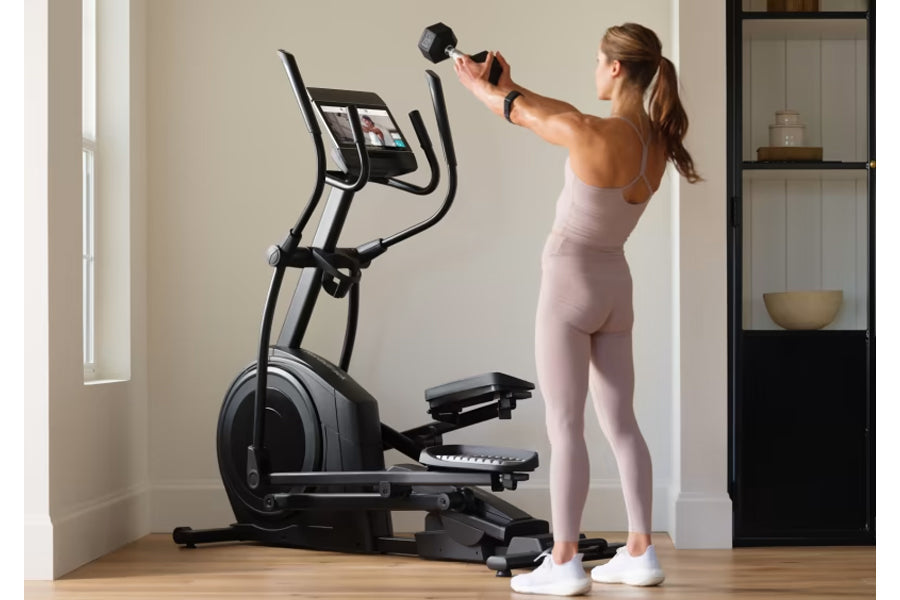 Woman wearing light pink workout set exercising on black NordicTrack elliptical machine with digital display in home gym