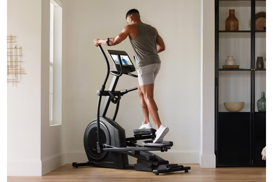Man wearing gray tank top and white shorts using black NordicTrack elliptical machine with digital display in home gym settin