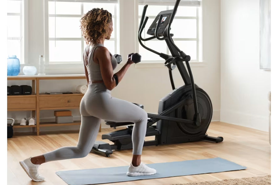 Woman performing lunge exercise with dumbbells on blue mat in front of black NordicTrack elliptical machine in home gym