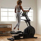 Woman wearing gray crop top and leggings using black elliptical machine in bright home gym with wooden furniture