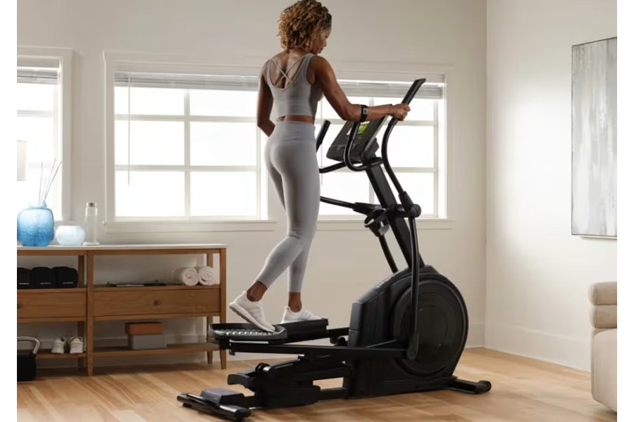 Woman wearing gray crop top and leggings using black elliptical machine in bright home gym with wooden furniture