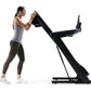 Woman using black NordicTrack treadmill, wearing gray tank top and brown leggings, demonstrated on white background