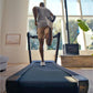Model running on NordicTrack Commercial 1750 treadmill in bright home gym with large windows, wearing gray tank top and tan s