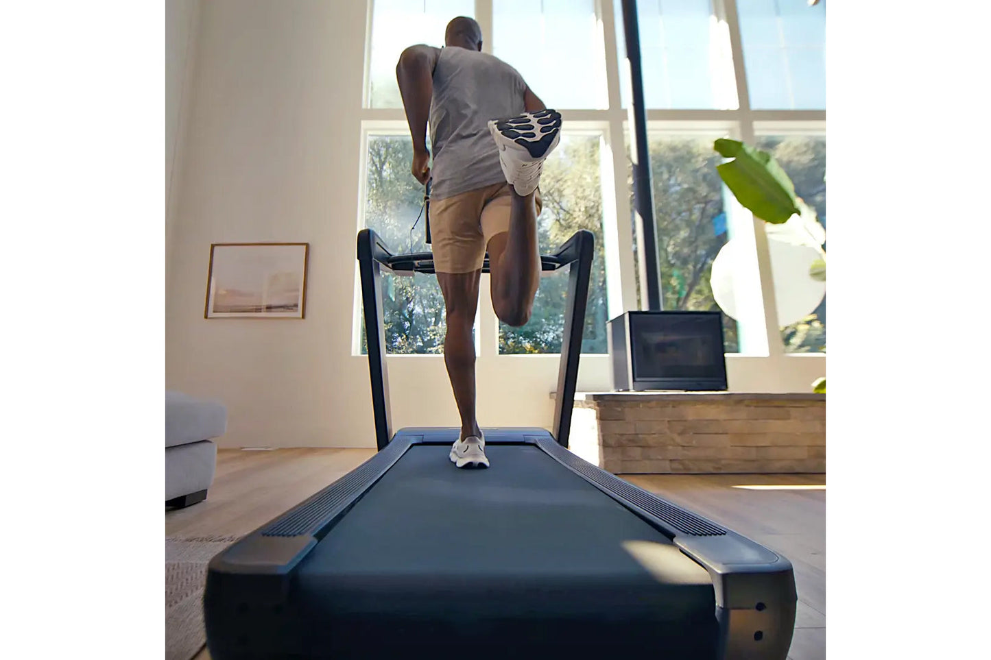 Model running on NordicTrack Commercial 1750 treadmill in bright home gym with large windows, wearing gray tank top and tan s