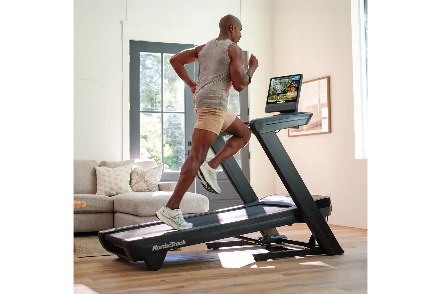Man running on black NordicTrack treadmill with tablet display in bright home gym with couch and windows