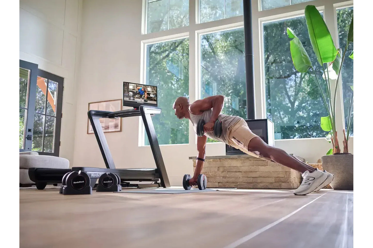 Man performing plank exercise on floor in modern home gym with NordicTrack treadmill, dumbbells, and potted plant visible