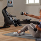 Woman with red curly hair using black NordicTrack rowing machine with attached screen, holding dumbbells on gray exercise mat