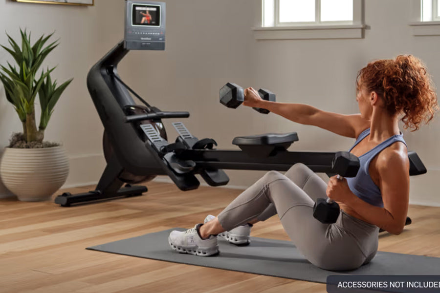 Woman with red curly hair using black NordicTrack rowing machine with attached screen, holding dumbbells on gray exercise mat