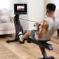 Black rowing machine with digital monitor displaying workout video, used by man in light gray tank top in living room