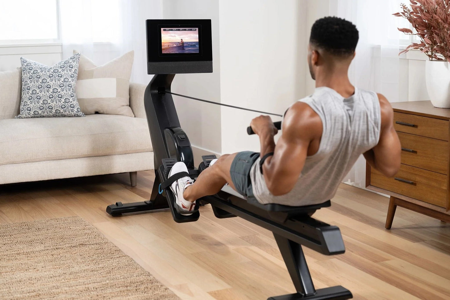 Black rowing machine with digital monitor displaying workout video, used by man in light gray tank top in living room