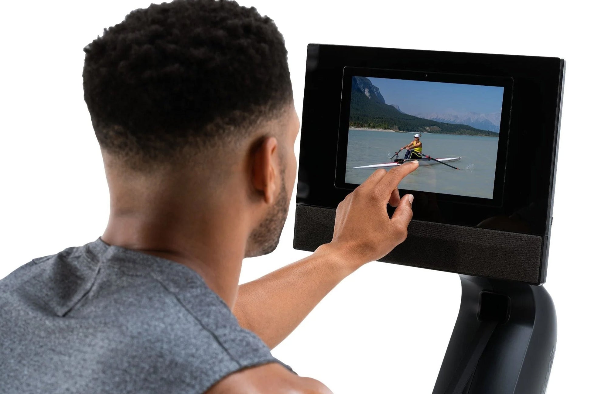 Man wearing gray shirt viewing black rower display screen showing rowing video on white background