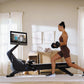 Woman in brown sports bra and shorts using black NordicTrack rowing machine with attached tablet screen in bright home gym