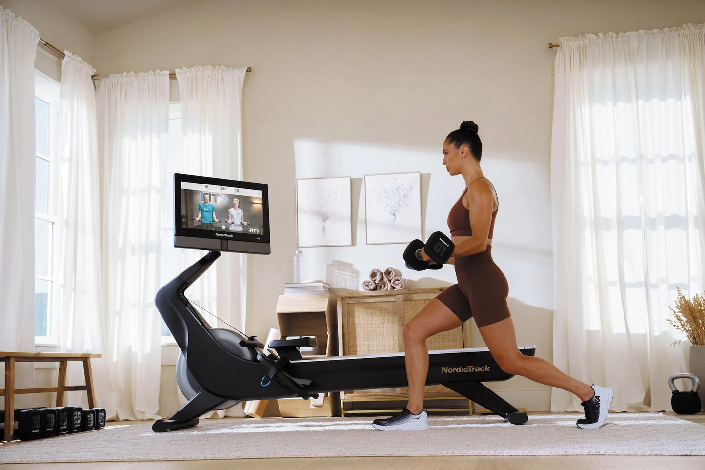 Woman in brown sports bra and shorts using black NordicTrack rowing machine with attached tablet screen in bright home gym