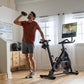Man in burgundy t-shirt and gray shorts exercising on black NordicTrack stationary bike while drinking water in home gym