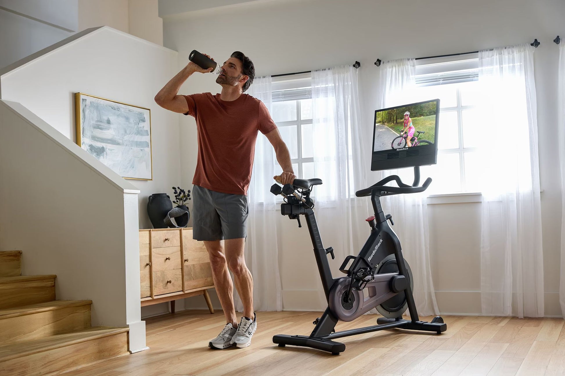 Man in burgundy t-shirt and gray shorts exercising on black NordicTrack stationary bike while drinking water in home gym
