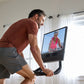 Man in burgundy t-shirt and gray shorts riding NordicTrack stationary bike with screen display showing cycling instructor