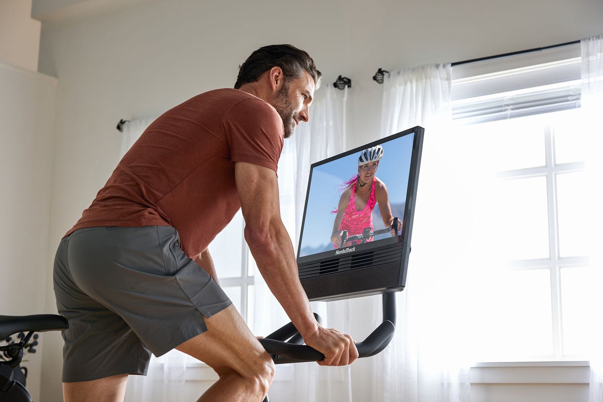 Man in burgundy t-shirt and gray shorts riding NordicTrack stationary bike with screen display showing cycling instructor