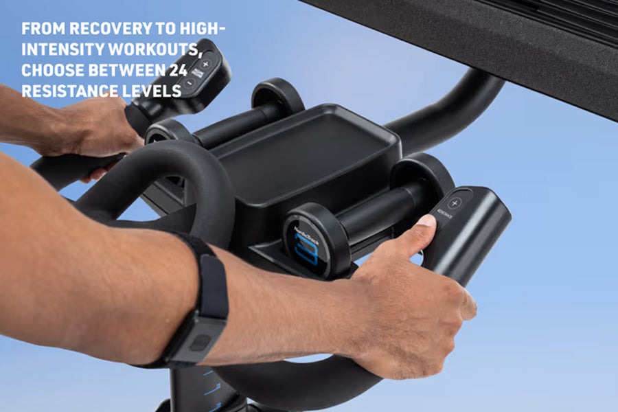 Black stationary exercise bike with digital display and resistance controls, held by person's hands against blue sky backgrou