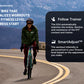 Woman in green jacket and burgundy pants riding teal indoor exercise bike on mountain road with snow-capped peaks in backgrou
