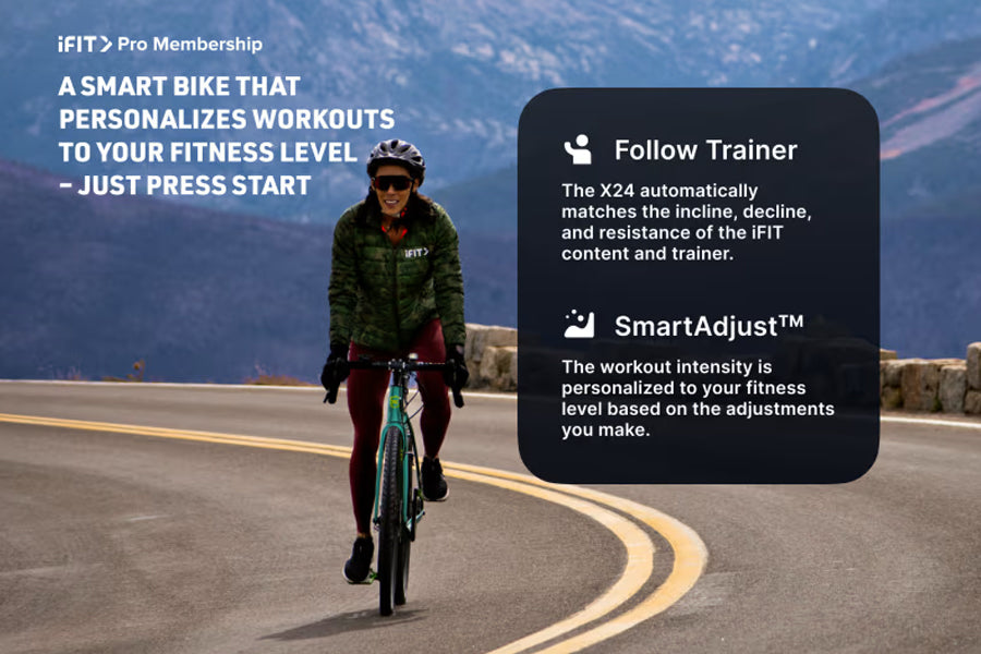 Woman in green jacket and burgundy pants riding teal indoor exercise bike on mountain road with snow-capped peaks in backgrou
