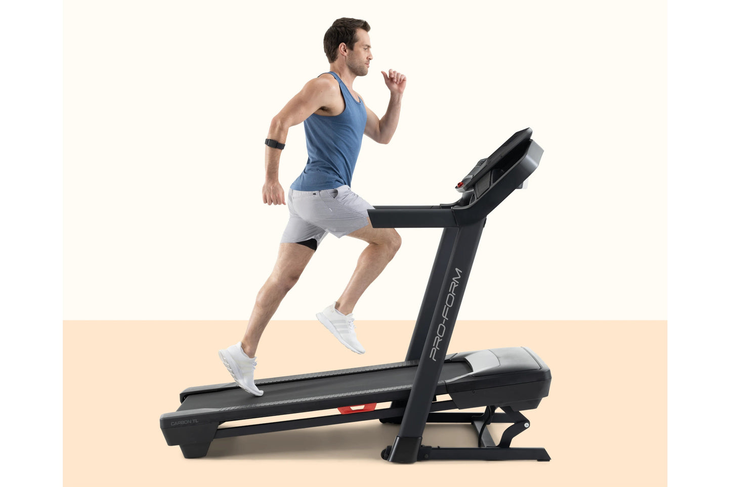 Man running on black ProForm Carbon TLS treadmill wearing blue tank top and white shorts, shown in profile on beige backgroun
