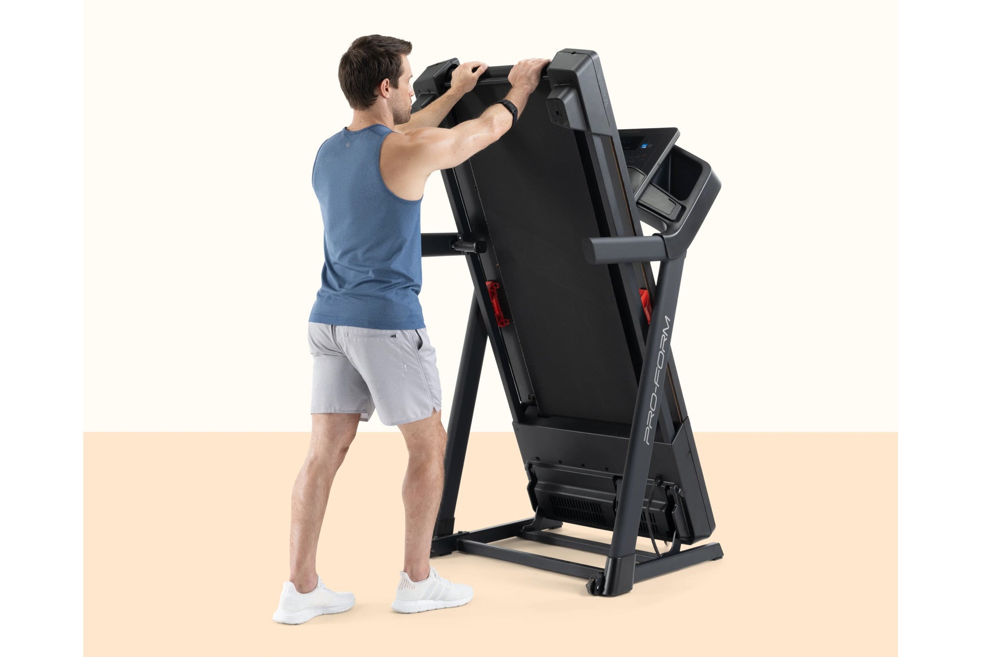 Man using black ProForm treadmill with digital display, wearing blue tank top and gray shorts on beige background