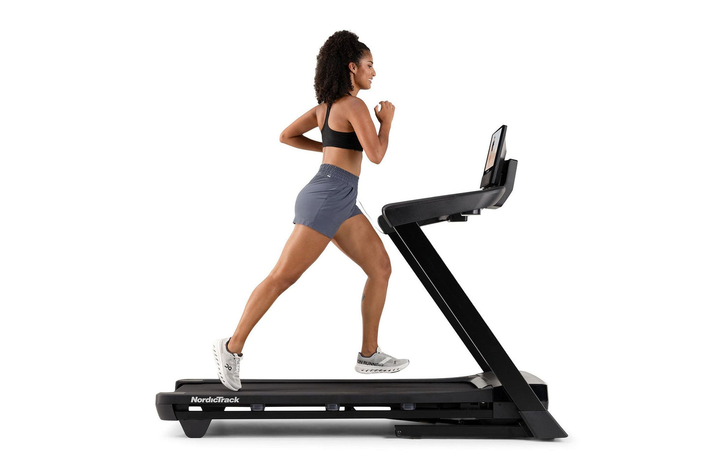 Woman running on NordicTrack black treadmill wearing black sports bra and gray shorts, white sneakers, on white background