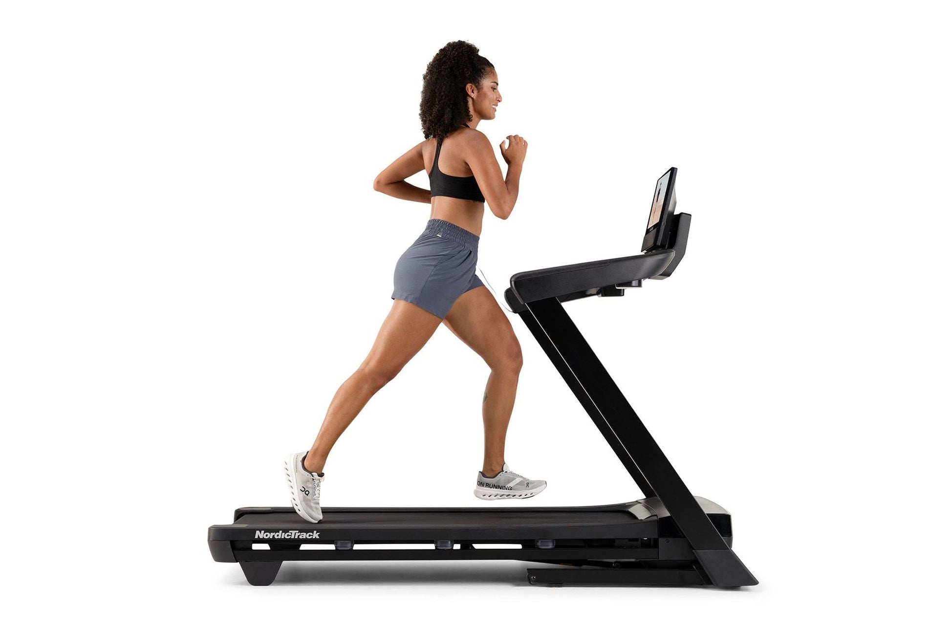 Woman running on NordicTrack black treadmill wearing black sports bra and gray shorts, white sneakers, on white background